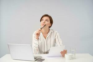 Puzzled young attractive brown haired woman with short trendy haircut keeping paper and pen in raised hands while looking pensively upwards, posing over white background photo