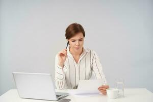 Concentrated young pretty short haired brunette woman holding pen in raised hand and looking at piece of paper with concerned face, biting worringly her lips while preparing report photo