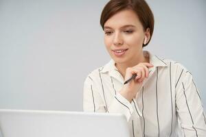 Portrait of beautiful young short haired brunette lady with trendy haircut raising hand with pen in it and smiling positively while looking on screen of her laptop, posing over white background photo