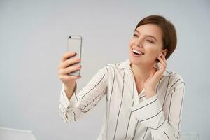 Indoor shot of young petty brown haired female with short trendy haircut keeping smartphone in raised hand while making video call, smiling broadly while posing over white background photo