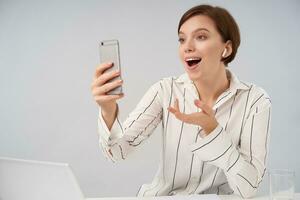 Excited young positive short haired brunette lady with casual hairstyle raising emotionally her palm and smiling widely while having video conversation with her mobile phone photo