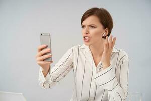 Displeased young pretty short haired brunette female frowning her eyebrows and looking at screen with pout while having stressful phone conversation, posing over white background photo