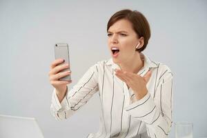 Stressed young brown-eyed short haired lady with natural makeup raising emotionally palm while having unpleasant video conversation with her smartphone, sitting over white background photo