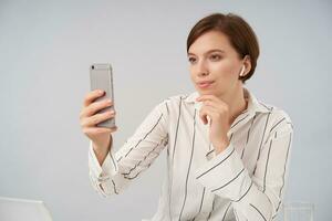 Good looking young positive short haired brunette lady dressed in elegant formal clothes making photo of herself with smartphone, holding her chin with raised hand while sitting over white background