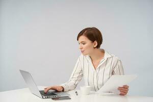 Indoor shor of lovely young brown haired lady with casual hairstyle working with modern laptop and looking attentively at screen, keeping documents in hand while posing over white background photo