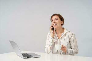 Indoor shot of pretty young brown haired woman with short trendy haircut keeping ceramic cup in raised hand while having pleasant talk on phone, sitting over white background photo