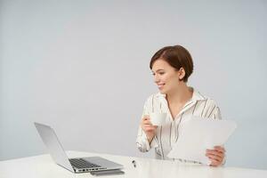 Positive young pretty brunette business woman with short trendy haircut holding cup of coffee and looking cheerfully at screen of her laptop, dressed in elegant formal clothes over white background photo