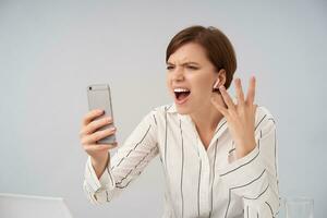 Emotional young pretty short haired brunette female with casual hairstyle frowning her face while shouting angrily over stressful video call, holding mobile phone while posing over white background photo