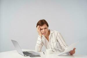 Displeased young brown haired female with natural makeup holding her head with raised hand and frowning her face while looking at documents, having a lot of work while sitting over white background photo