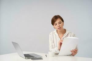 Beautiful young short haired brunette woman with natural makeup preparing materials for upcoming meeting, sitting over white background with cup of tea and laptop photo