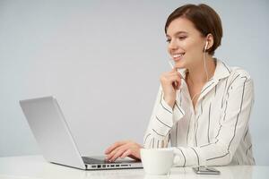 Shot of attractive young brown haired woman with short trendy haircut holding hand on keyboard while sitting over pink background with laptop, smiling happily and holding raised hand under her chin photo