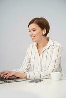 Positive young lovely short haired brunette lady listening to music in her headphones while typing text with keyboard on laptop, sitting over white background in striped shirt photo