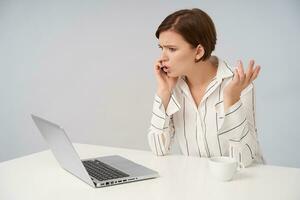 Confused young pretty short haired brunette female with casual hairstyle raising her palm while solving problems during phone call, frowning her face while looking in front of herself photo