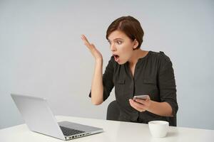 Shocked young short haired brunette woman with casual hairstyle raising emotionally palm and looking at her laptop with wide eyes and mouth opened, isolated over white background photo