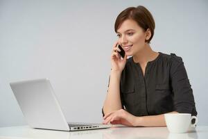 Positive young pretty short haired brunette female dressed in black shirt working at modern office with laptop and making call with her samrtphone, looking cheerfully on screen photo