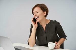 Joyful young lovely short haired brunette woman with natural makeup making break with work and calling her friend, laughing happily while sitting over white background with ceramic cup in her hand photo