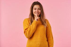 Indoor shot of young lovely brown haired woman with casual hairstyle keeping forefinger on her lips while looking positively at camera, asking to keep secret, isolated over pink background photo
