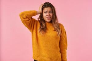 Bemused young attractive brunette woman raising hand to her head while looking confusedly at camera, frowning eyebrows and wrinkling forehead while posing over pink background photo