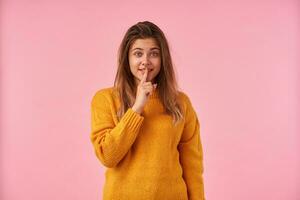 Studio photo of young pretty brown haired lady with casual hairstyle holding index finger on her lips while standing over pink background, asking not to breathe a word about news she told