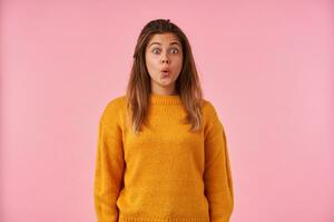 Amazed young brown haired woman with natural makeup raising surprisedly eyebrows while looking at camera with round eyes, standing against pink background photo