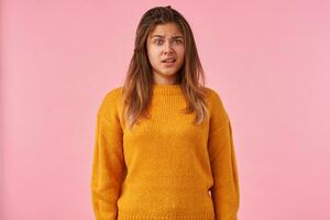 Puzzled young attractive brown haired woman raising wonderingly eyebrow and wrinkling her forehead while posing over pink background in warm knitted mustard sweater photo