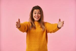 Cheerful young attractive brown haired lady raising happily eyebrows and smiling broadly while standing over pink background with opened arms, dressed in mustard warm sweater photo