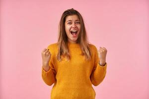 Overjoyed young pretty brunette lady with natural makeup screaming happily with wide mouth opened and raising her fists, wearing casual clothes while posing over pink background photo