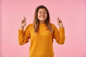 Pleasant looking hoping young lovely brunette female keeping her eyes closed while crossing fingers and making wish, biting underlip while standing over pink background photo