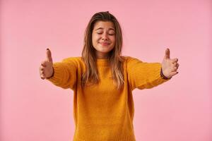 Pleasant looking young brown haired woman keeping her eyes closed while smiling positively, spreading her raised hands while posing over pink background in casual clothes photo