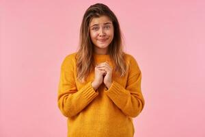 Studio shot of hoping lovely young brown haired lady pursing her lips and looking desperately at camera, keeping raised hands together while posing over pink background photo