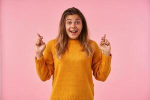 Overjoyed young attractive brown haired woman raising excitedly her eyebrows while looking cheerfully at camera and crossing her fingers for good luck, isolated over pink background photo