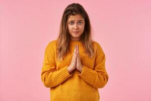 Desperate young pretty brown haired woman looking sadly at camera with folded lips, raising palms in praying gesture while standing against pink background photo