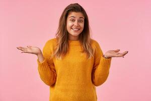 Studio photo of young lovely brunette woman with casual hairstyle shrugging with raised hands and looking positively at camera with embarrassed smile, isolated over pink background