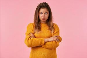 Indoor shot of offended young brown haired pretty woman in mustard warm sweater folding her hands while posing over pink background, looking at camera with displeased face and pouting lips photo