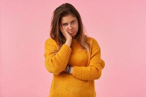 Upset young brown haired female with natural makeup leaning her head on raised hand and looking sadly to camera with pursed lips, isolated over pink background photo