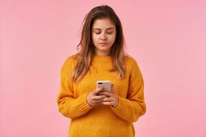 Sudio shot of young brown haired lady with natural makeup holding smartphone while standing over pink background, looking attentively at screen and keeping her lips folded photo