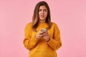Puzzled young pretty brown haired lady with casual hairstyle holding mobile phone and looking at camera with confused face, dressed in mustard knitted sweater over pink background photo