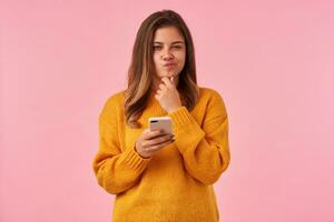 Pensive young brown haired woman with natural makeup holding her chin with raised hand and squinting thoughtfully while looking at camera with pouted lips, isolated over pink background photo