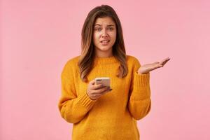 Indoor shot of young brown haired confused female looking at camera with displeased face and raising confusedly hand while standing against pink background with smartphone photo