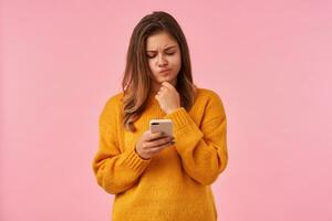 Bewildered young pretty brown haired female woman twisting her mouth and frowning confusedly eyebrows while looking at screen on her phone, isolated over pink background photo