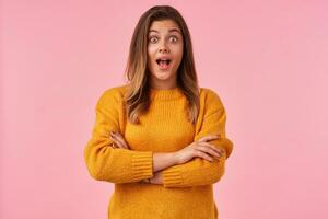 Overjoyed young brown haired pretty female looking excitedly at camera with wide eyes and mouth opened, dressed in casual clothes while standing over pink background photo