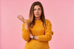 Dissatisfied young pretty brown haired woman dressed in mustard knitted sweater rolling her eyes and raising hand discontentedly, standing against pink background photo