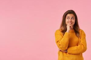 Studio photo of positive lovely young brown haired woman with natural makeup holding her chin and looking dreamily upwards, wearing mustard knitted sweater over pink background