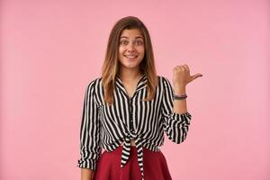 Indoor shot of charming young brunette woman with natural makeup pointing aside with thumb and looking positively at camera with pleasant smile, isolated over pink background photo