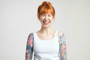 Joyful young attractive redhead woman with tattoos keeping her eyes closed while laughing cheerfully, dressed in white shirt while posing over white background photo