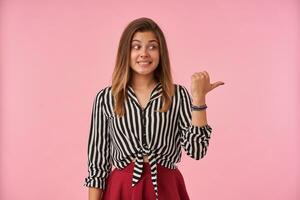 Puzzled young attractive brown haired lady with casual hairstyle showing her perfect white teeth while looking confusedly aside, standing against pink background photo