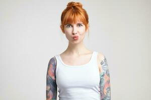 Horizontal shot of young lovely tattooed female with bun hairstyle pouting her lips while fooling, dressed in casual wear while posing over white background photo