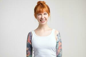 Studio photo of young lovely redhead woman with tattoos squinting her eyes while smiling joyfully, being in high spirit while posing over white background