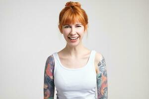 Joyful young attractive tattooed woman with foxy hair laughing happily while looking cheerfully at camera, standing over white background with hands down photo