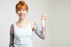 Indoor shot of young attractive redhead woman with tattoos frowning her face while smiling and thumbing aside with raised hand, isolated over white background photo